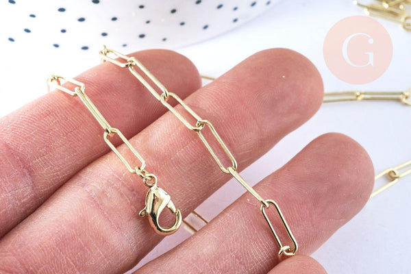 Close-up of fingers holding a gold paper-clip chain necklace with rectangular links and a lobster-claw clasp. Product: Collier Chaine laiton doré clair maille rectangle.