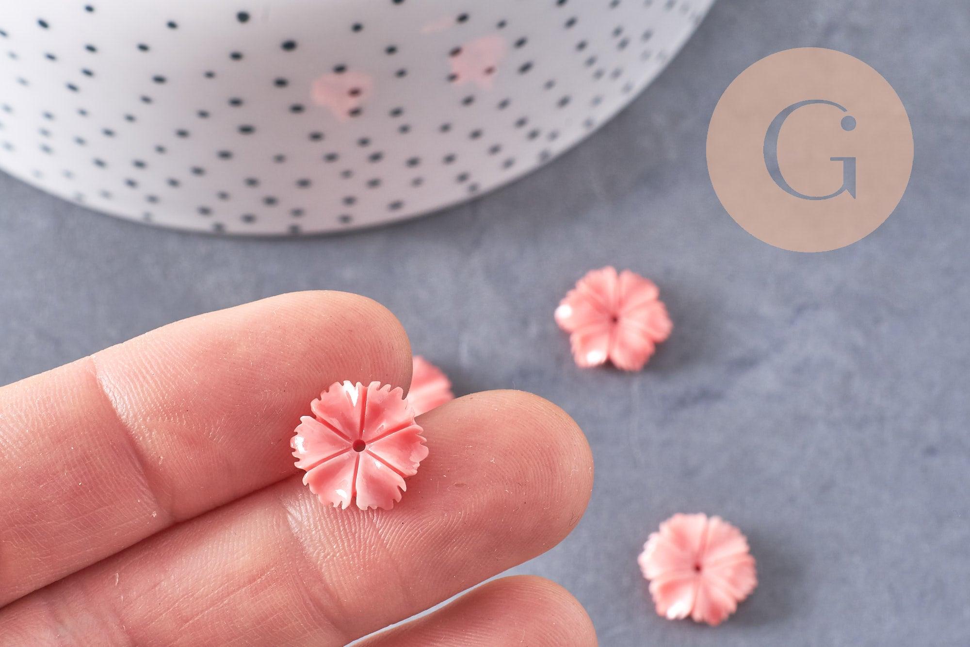 Close-up of fingertips holding a pink, flower-shaped resin bead, 13mm, with similar beads scattered below; product: EP-Perle fleur résine rose, x5, for jewelry making.
