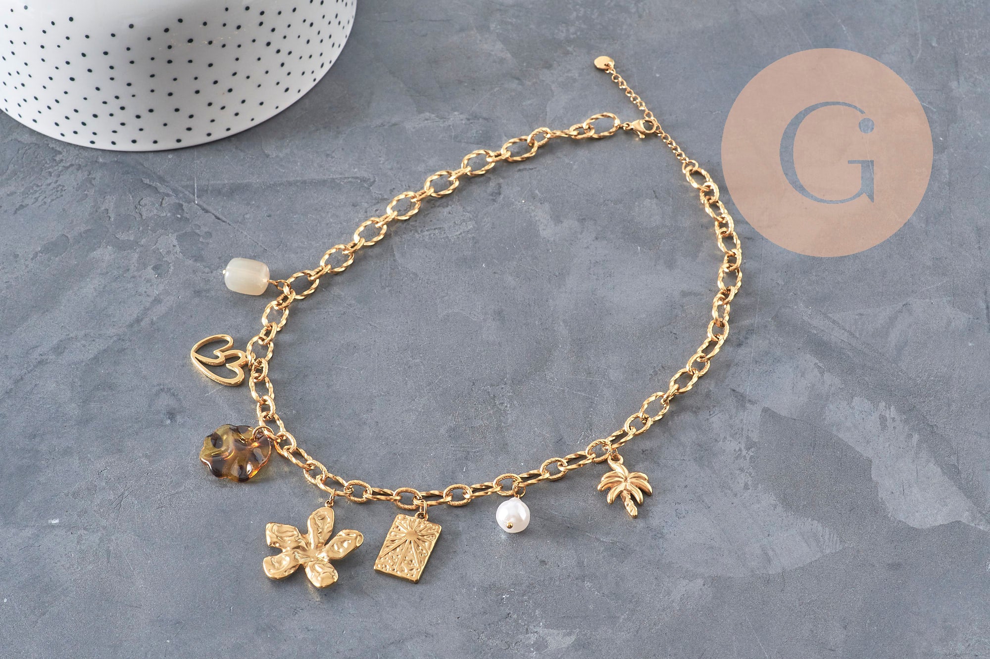 Collier doré en acier inoxydable avec chaîne à maillons et breloques variées, perles et médaillons sur fond gris.