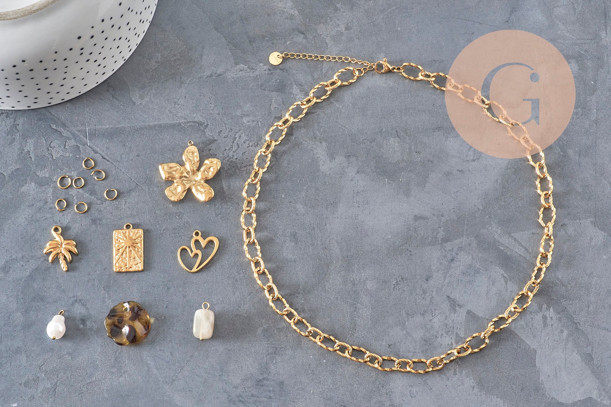 Kit collier DIY avec chaîne en acier inoxydable doré, breloques fleur, cœur, arbre de vie et perles sur fond gris.
