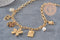 Collier DIY en chaîne dorée acier inoxydable 304 avec breloques cœur, fleur, perles nacrées et médaillons sur fond gris.
