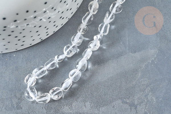 Translucent pebble-shaped glass beads, Perles cristal de roche roulée pierre brute, lie on a dark gray stone surface, beside a dotted white ceramic pot.